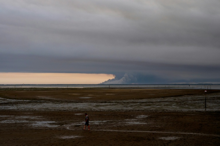 Un homme marche sur une plage d'Andernos au bord du bassin d'Arcachon, au fond duquel s'élèvent les fumées de l'incendie de Biscarosse, le 23 juillet 2026