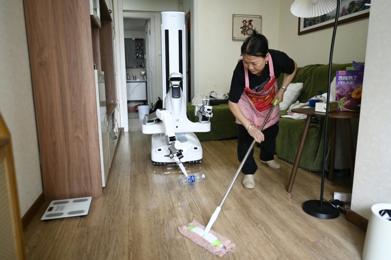 Un robot assistant ménager transporte une bouteille jusqu'à une poubelle tandis qu'une femme de ménage nettoie le sol chez une cliente à Pékin, le 21 mai 2026 en Chine