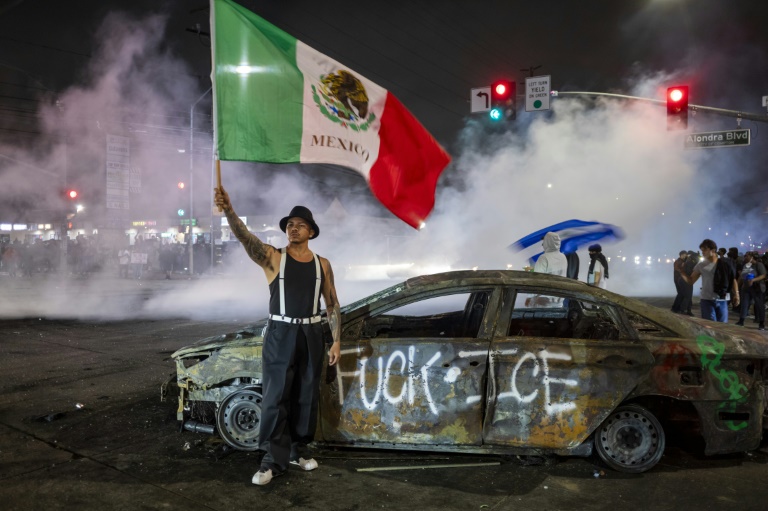 Un manifestant tient un drapeau mexicain devant l'épave d'une voiture incendiée lors de heurts avec les forces de l'ordre contre l'expulsion d'immigrés, à Los Angeles, le 7 juin 2025 en Californie